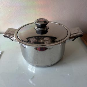 May include: Stainless steel cooking pot with a lid. The pot has two handles and a black knob on the lid. The pot is sitting on a white surface. The pot is made of shiny metal.