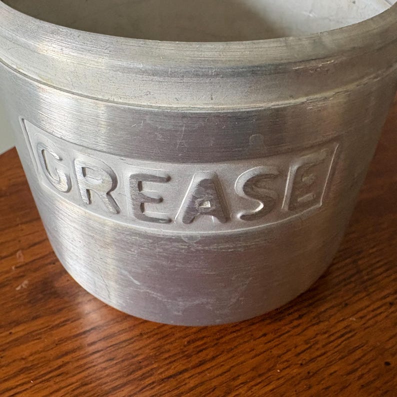 May include: A vintage aluminum grease container with the word "GREASE" embossed on the side. The container has a cylindrical shape with a slightly flared rim. The metal has a brushed finish, showing signs of wear.
