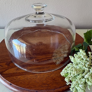 May include: A clear glass cake dome with a round wooden base. The dome has a small handle on top. The wooden base is a rich, dark brown color. The dome is sitting on a round, wooden base. A sprig of white flowers and green leaves are on the right side.