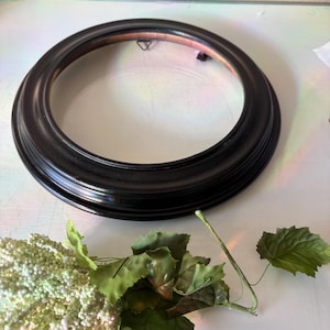 May include: A round, black picture frame with a decorative edge. The frame is empty, with a clear glass or plastic insert. A sprig of green leaves and white flowers is in the foreground.