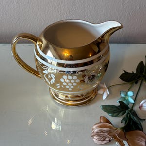 May include: A gold-toned ceramic creamer pitcher featuring a white interior and a glossy metallic exterior finish. The body is decorated with horizontal white bands and a white grape cluster pattern. It has a curved handle and a pouring spout.