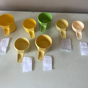 May include: A collection of yellow and green measuring cups of various sizes. The cups are arranged on a white surface, with handwritten labels indicating measurements. The cups are made of plastic and have handles.