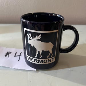 May include: A dark blue ceramic mug with a white moose silhouette and the word "VERMONT" in a white rectangle. The mug has a black handle and a glossy finish. A piece of paper with the number "4" is in the foreground.