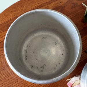May include: A vintage, round, aluminum pot with a wide rim. The interior shows signs of use with dark spots. The pot sits on a wooden surface. This cooking pot is likely used for preparing food.