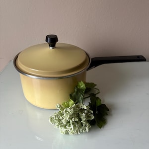 May include: A vintage yellow saucepan with a black handle and lid. The pot has a silver rim and a black knob on the lid. Green and white faux flowers are placed in front of the pot.