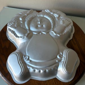 May include: A silver-colored metal cake pan in the shape of a doll. The pan features the doll's face, hair, arms, legs, and clothing details. The doll has a smiling expression and is wearing a dress. The pan is designed for baking a cake in the shape of the doll.