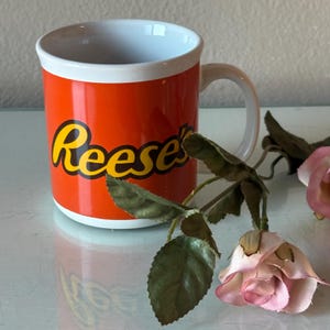 May include: An orange ceramic mug with a white interior and handle. The mug features the word "Reese's" in yellow script. A pink rose and green leaves are in the foreground.