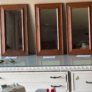 May include: Three rectangular mirrors with brown wooden frames are displayed on a white dresser. The mirrors reflect the room, and the frames have a simple, classic design. The dresser has white drawers with black handles.