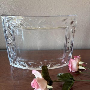 May include: A clear, curved glass picture frame featuring an etched leaf and vine pattern along its border. The frame has a rectangular center opening and a smooth, polished finish, showcasing a transparent design with intricate detailing.