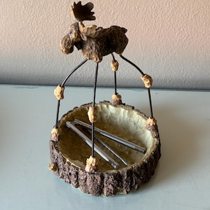 May include: A whimsical decorative bowl featuring a carved wooden moose figure with long legs. The bowl is made to look like a tree trunk and contains silver nutcrackers. The moose is brown and the bowl has a light beige interior.