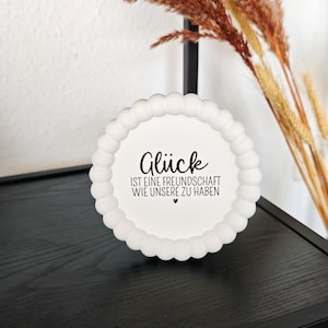 Könnte beinhalten: Eine weiße, dekorative Platte mit gewelltem Rand und dem deutschen Text "Glück ist eine Freundschaft wie unsere zu haben" in Schwarz. Die Platte steht auf einer schwarzen Oberfläche, mit getrockneten Ziergräsern im Hintergrund.