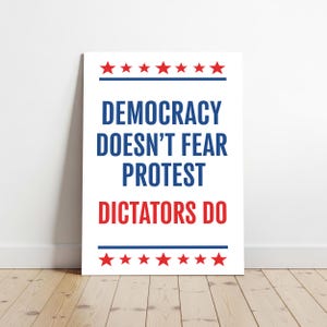 May include: White poster with the text "DEMOCRACY DOESN'T FEAR PROTEST DICTATORS DO" in blue and red. Red stars are at the top and bottom of the poster. The poster is leaning against a white wall on a wooden floor.