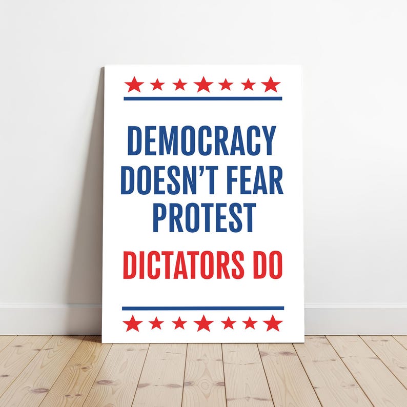May include: White poster with the text "DEMOCRACY DOESN'T FEAR PROTEST DICTATORS DO" in blue and red. Red stars are at the top and bottom of the poster. The poster is leaning against a white wall on a wooden floor.