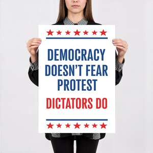 May include: A white poster with the text "DEMOCRACY DOESN'T FEAR PROTEST DICTATORS DO" in blue and red. The poster has red stars and blue lines at the top and bottom. The poster is held by a person.