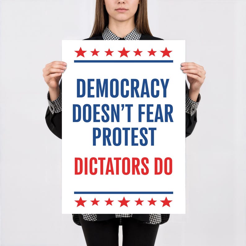 May include: A white poster with the text "DEMOCRACY DOESN'T FEAR PROTEST DICTATORS DO" in blue and red. The poster has red stars and blue lines at the top and bottom. The poster is held by a person.