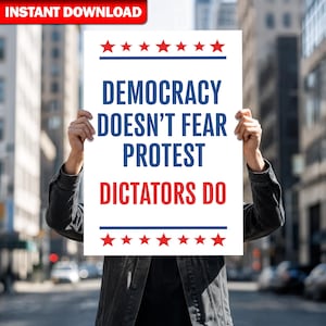 May include: A white poster with the text "DEMOCRACY DOESN'T FEAR PROTEST DICTATORS DO" in blue and red, with red stars and blue lines. The poster is held up in front of a person in a black jacket.