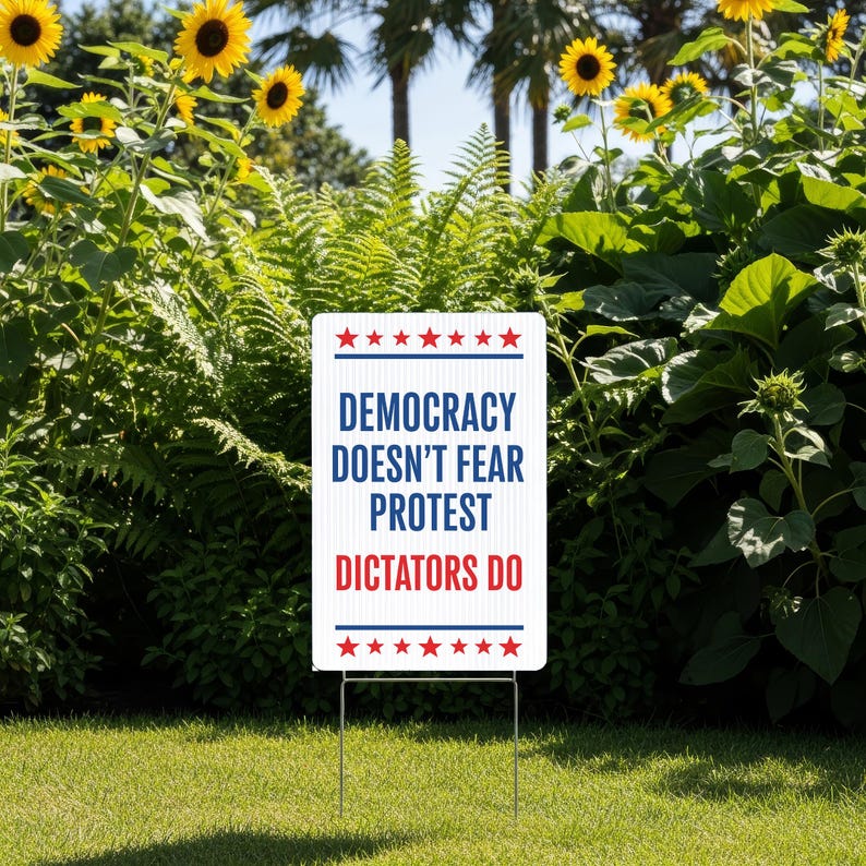 May include: A white rectangular yard sign with the text "DEMOCRACY DOESN'T FEAR PROTEST DICTATORS DO" in blue and red. The sign is supported by a metal stake and is placed in a grassy area with sunflowers and greenery in the background.