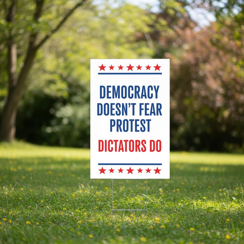 May include: A white yard sign with the text "DEMOCRACY DOESN'T FEAR PROTEST DICTATORS DO" in blue and red. The sign has red stars and blue stripes at the top and bottom. It is placed in a grassy yard.