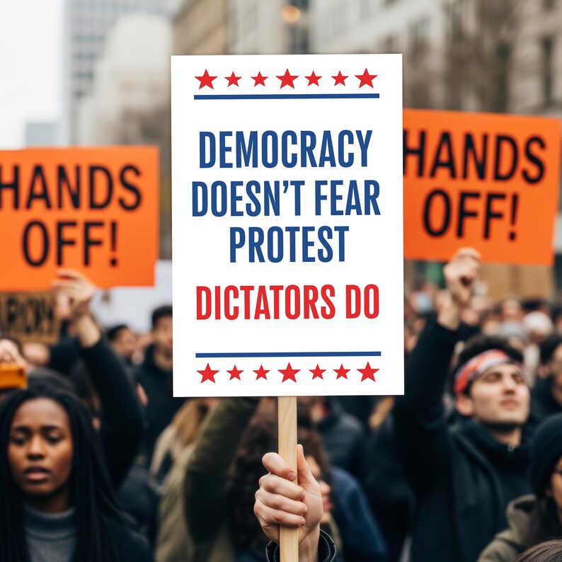 May include: A protest sign held high, with the words "DEMOCRACY DOESN'T FEAR PROTEST DICTATORS DO" in blue and red, framed by stars. Other signs read "HANDS OFF!" in orange. A crowd of people is visible in the background.