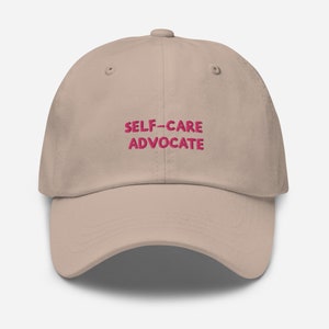 May include: A beige baseball cap featuring the embroidered text SELF-CARE ADVOCATE in a bold pink font centered on the front panel. The cap has a structured crown, a curved brim, and matching stitching details across the fabric surface.