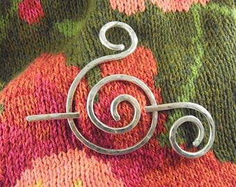 May include: Silver spiral shawl pin with a hammered finish. The pin is on a red and green knitted fabric.
