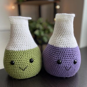 May include: Two handmade crochet flasks, one green and one purple, each with a white top. The flasks have black button eyes and stitched smiles. These whimsical, decorative items are perfect for a science-themed nursery or home decor.