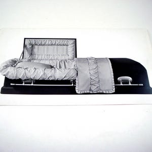 May include: A black casket with a silver interior, open to display the interior lining and pillow. The casket is on a white background. The interior is a light gray color with a pleated design.