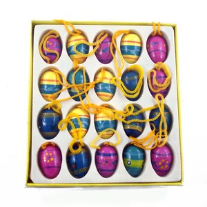 May include: A set of fifteen painted wooden Easter egg ornaments in a yellow box. The eggs are oval-shaped with various colors, including blue, yellow, and pink, and are strung with yellow cords. The box is white with a yellow border.