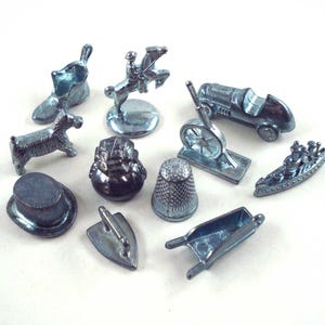 May include: A collection of silver-colored Monopoly game tokens. The tokens include a shoe, a horse and rider, a race car, a dog, a top hat, a thimble, a wheelbarrow, an iron, a battleship, and a cannon.