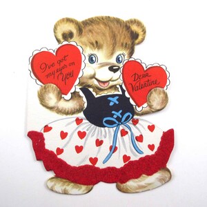 May include: A vintage Valentine's Day card featuring a cartoon teddy bear wearing a white dress with red hearts, a blue apron, and holding two red hearts. The hearts have the text "I've got my eyes on you" and "Dear Valentine".
