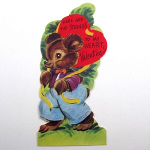 May include: Vintage Valentine's Day card featuring a brown bear wearing a purple jacket and blue pants. The bear holds a red heart with the text "Here are the strings to my heart, Valentine." The card has a green leafy background and yellow ribbons.