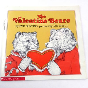 May include: A children's book cover titled "The Valentine Bears" by Eve Bunting, with illustrations by Jan Brett. The cover features two bears holding a red heart. The bears are wearing red jackets. The background is yellow.