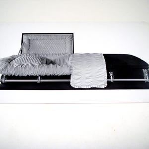 May include: A black casket with a silver interior, including a pillow and a blanket. The casket is open, revealing the interior lining. The image is a black and white photograph.