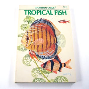 May include: A softcover book titled A Golden Guide Tropical Fish featuring a large orange and brown discus fish, a smaller orange and black striped fish, and a tiny red fish swimming among green aquatic plants on a cream-colored background.