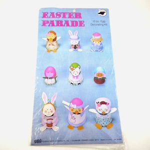 以下が含まれることがあります： 「Easter Parade」の文字が入った、10個入りのイースターエッグデコレーションキット。ウサギ、ヒヨコ、その他のフィギュアなど、さまざまな卵型のキャラクターが特徴です。「10 pc. Egg Decorating Kit」の文字も表示されています。