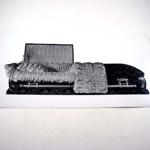 May include: A black and silver casket with an open lid, revealing a patterned interior. A decorative silver blanket rests on the closed portion. The casket has silver handles and is set against a white background.