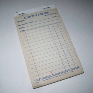 May include: A white paper receipt book with blue lines and the text "Scotty's Korner" printed in black. The receipt book has the address "West Main St. Princeton, Ky. 42443" and the phone number "365-6236" printed on it. The bottom of the receipt book has the text "Bank Americard, Master Charge & Layaway" printed in red and black. The text "Lakewood Printers, Princeton, Ky. 42443" is printed in black.