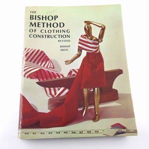 May include: The cover of a vintage book titled "The Bishop Method of Clothing Construction Revised" by Bishop Arch. The cover features a wooden dress form wearing a red and white striped top and a red skirt. The dress form is standing on a wooden table with a red and white striped fabric draped over it.