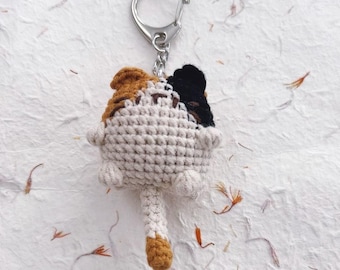 May include: A crocheted keychain featuring a small, round, cream-colored cat figure with a calico pattern. The cat has a brown patch on the left ear, a black patch on the right ear, and a long, cream-colored tail with a brown tip attached to the bottom.