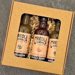 May include: A gift box containing three glass bottles of pepper sauce. The bottles have brown labels with the text "REOLA TRADING CO" and "CREOLA TRADING CO". The box is made of brown cardboard and filled with shredded paper.