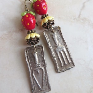 May include: A pair of dangle earrings featuring red strawberry beads with green leaves and yellow dots. Below the strawberries are cream-colored flower beads and rectangular silver-tone charms with a garden tool and heart design. The earrings have brass-colored ear wires.