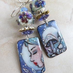 May include: Handmade dangle earrings with a celestial theme. Each earring features a rectangular pendant with a blue background and a painted design of a woman's face and a mushroom. The earrings also have floral and bead accents.