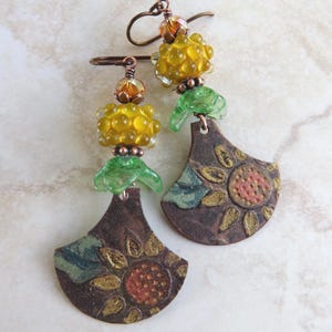 May include: A pair of dangle earrings featuring copper-toned metal fan-shaped charms embossed with sunflower designs. Each earring includes a translucent green leaf-shaped bead, a textured yellow glass bead, and a small amber crystal bead.