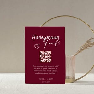 Puede incluir: Una tarjeta burdeos con las palabras "Honeymoon Fund" en escritura blanca, un símbolo de corazón y un código QR. La tarjeta incluye texto sobre la contribución a un fondo de luna de miel. Los nombres Olivia y Aaron, y la fecha 20.09.2027 también están impresos.