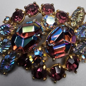 May include: Vintage brooch featuring a floral design with iridescent and purple gemstones set in a gold-tone metal. The brooch has large, faceted center stones and smaller, round and leaf-shaped accents.