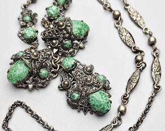May include: An antique silver-toned necklace and matching earrings set. The necklace features a chain with floral and oval links, and the earrings have large, oval green speckled stones. The set has intricate filigree detailing.