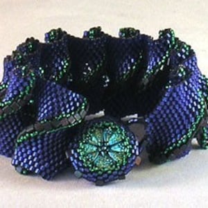 Puede incluir: Un brazalete de cuentas azul oscuro y verde con un bot&oacute;n grande de color verde en el centro. El brazalete est&aacute; hecho de cuentas peque&ntilde;as, tejidas apretadamente y tiene un dise&ntilde;o con volantes y textura.