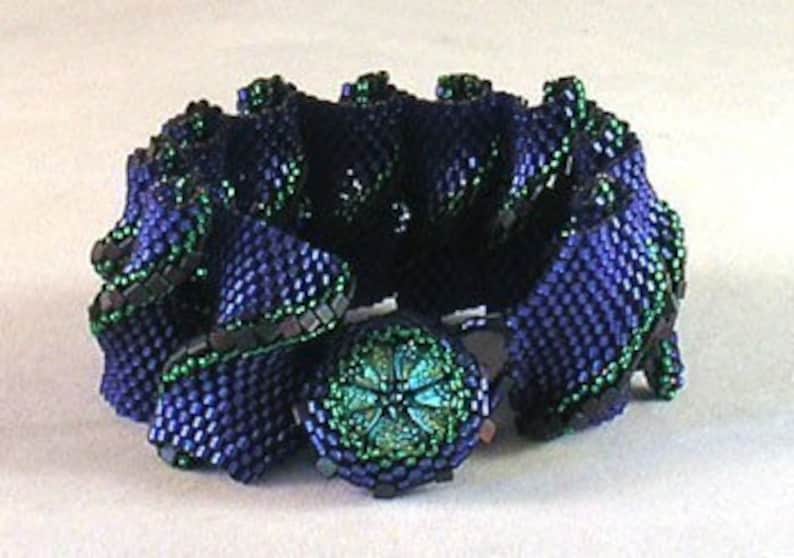 Puede incluir: Un brazalete de cuentas azul oscuro y verde con un bot&oacute;n grande de color verde en el centro. El brazalete est&aacute; hecho de cuentas peque&ntilde;as, tejidas apretadamente y tiene un dise&ntilde;o con volantes y textura.