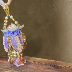 May include: A detailed, handmade ornament with a beaded handle and a decorative tassel. The ornament is adorned with gold, pink, purple, and white beads and sequins. The design incorporates a floral element and a delicate tassel.