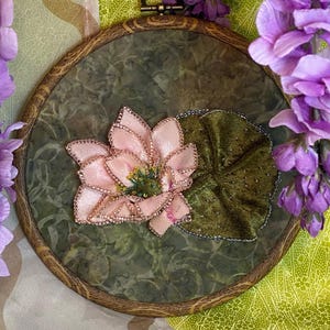 May include: A handcrafted embroidery hoop featuring a pink lotus flower with gold accents and a green velvet leaf. The artwork is set against a mottled green background within a wooden hoop, with purple flowers in the background.